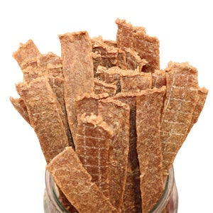 May include: A close-up of a jar filled with long, thin, brown, dog treats. The treats are stacked on top of each other and have a slightly textured surface.