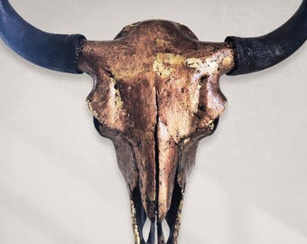 May include: A decorative bull skull with a metallic bronze finish and gold leaf accents. The horns are painted black. This rustic piece is suitable for wall decor and adds a touch of Western style to any space.
