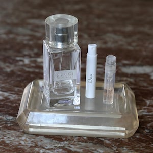 May include: A clear glass perfume bottle labeled "GUCCI Eau de Parfum II" stands on a rectangular tray, accompanied by two smaller perfume vials. One vial is white and labeled "Dior". The other vial is clear with a light pink liquid.