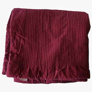 May include: A folded, burgundy-colored blanket with a textured, quilted design. The blanket features a braided pattern across the center and a smooth, velvet-like border. The fabric appears soft and luxurious, suitable for home decor.