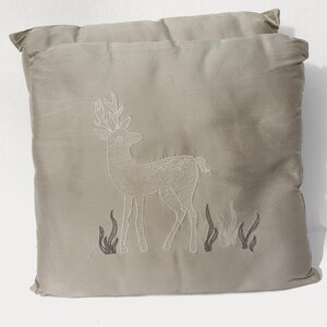 May include: A beige throw pillow with an embroidered white deer and black foliage design.