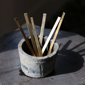 May include: A collection of brass herb markers, each labeled with a different herb name: oregano, jalapeno, rosemary, lemon balm, and basil. The markers are displayed in a small, weathered, gray container, set on a dark wooden surface.