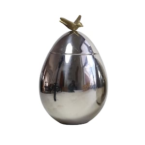 May include: A silver-coloured, egg-shaped container with a lid, topped with a small, gold-coloured bird figurine. The container has a reflective surface, and the bird is perched on top. The overall design is elegant and decorative.