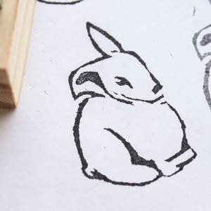 May include: A black and white line drawing of a rabbit sitting with its head tilted to the side. The rabbit is drawn in a simple, cartoon style.