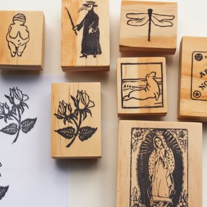 May include: A set of nine wooden stamps with various designs, including a woman with her arms crossed, a plague doctor, a dragonfly, a seal, a rose, and a Ouija board.