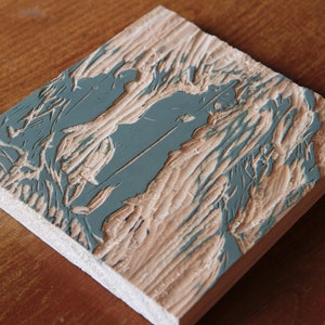 May include: A close-up of a wooden printing block with a carved design. The design is painted in a light blue color and features a stylized image of a tree with branches and leaves.