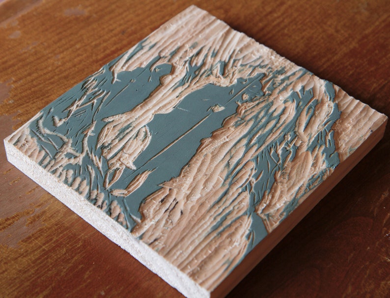 May include: A close-up of a wooden printing block with a carved design. The design is painted in a light blue color and features a stylized image of a tree with branches and leaves.