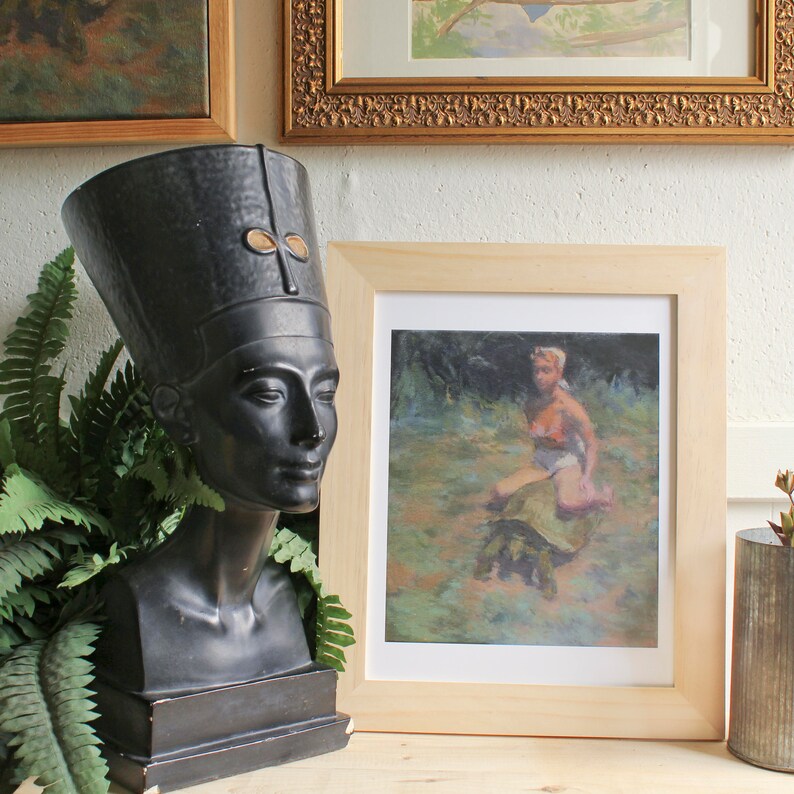 May include: A black bust sculpture of an Egyptian queen wearing a headdress with a gold symbol. The sculpture is on a wooden surface next to a framed picture of a woman sitting on a turtle in a forest setting.