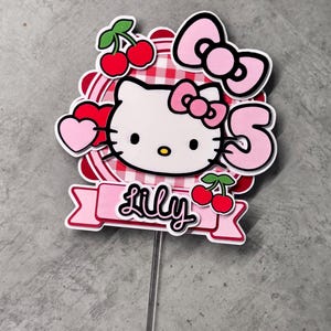 May include: A pink and red Hello Kitty cake topper with a red and white gingham background, cherries, and the name "Lily" written on a pink banner.