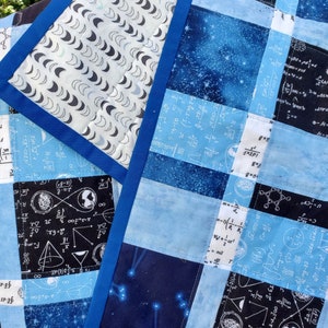 May include: A blue and white patchwork quilt with a variety of science-themed patterns, including constellations, mathematical equations, and scientific diagrams. The quilt is edged with a blue binding.