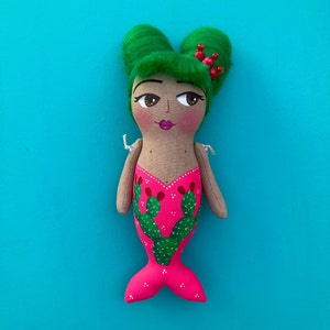 Puede incluir: Una muñeca hecha a mano con cabello verde y un vestido rosa decorado con diseños de cactus verdes. La muñeca tiene una cara pintada con ojos marrones y labios rojos.