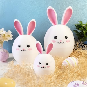 Peut inclure: Trois figurines de lapin de Pâques en céramique blanche avec des oreilles roses. Les lapins sont de tailles différentes et ont des yeux noirs et un nez noir. Les lapins sont assis sur un lit de papier déchiqueté.