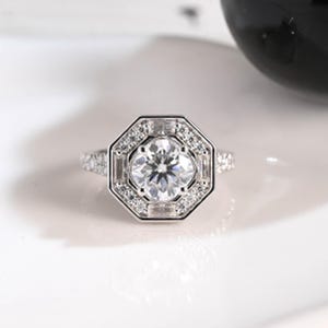 Puede incluir: Un anillo plateado con una piedra preciosa grande, redonda y transparente en el centro, rodeada de piedras más pequeñas y transparentes en un diseño geométrico. La banda del anillo también está adornada con pequeñas piedras transparentes. El anillo está sobre un fondo blanco.