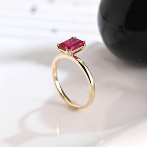 May include: A gold ring featuring a rectangular, emerald-cut vibrant red gemstone set in a four-prong setting. The band is thin, smooth, and polished, showcasing a minimalist design that highlights the deep crimson color of the central stone.