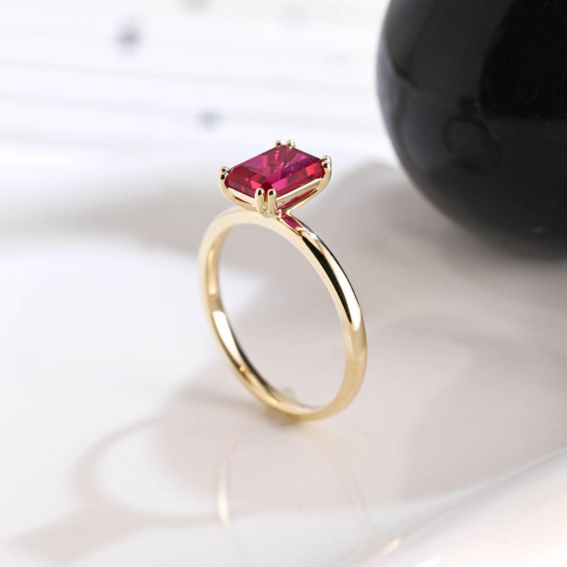May include: A gold ring featuring a rectangular, emerald-cut vibrant red gemstone set in a four-prong setting. The band is thin, smooth, and polished, showcasing a minimalist design that highlights the deep crimson color of the central stone.