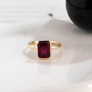 May include: A gold ring with a rectangular, deep red gemstone. The gemstone is set in a bezel setting, and the band is a simple, polished gold. The ring is displayed on a white surface.