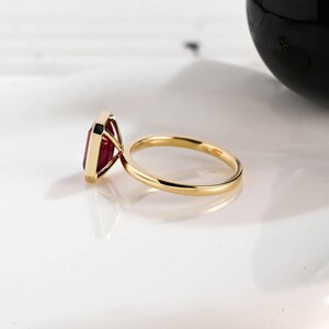 May include: A gold ring with a large, rectangular, deep red gemstone. The ring's band is thin and polished, and the gemstone is set in a gold bezel. The ring is displayed on a white surface.