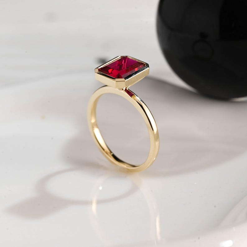 May include: A gold ring with a rectangular, deep red gemstone. The ring has a thin band and a bezel setting for the gemstone. The gemstone is a vibrant red color, and the ring is set against a white background.