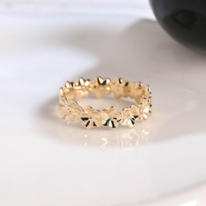 May include: A gold-colored ring featuring a repeating floral design. The ring is made of a shiny, reflective metal and sits on a white surface. The flowers are evenly spaced around the band, creating a continuous pattern.