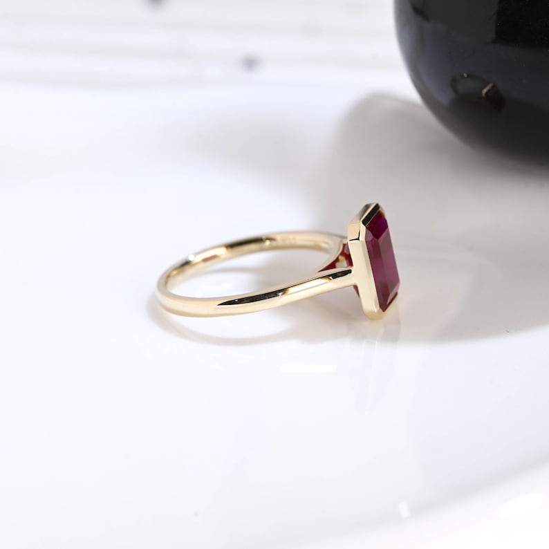 Puede incluir: Un anillo de oro con una piedra preciosa roja rectangular de corte esmeralda en un engaste de bisel. La banda es delgada y pulida, con un acabado met&aacute;lico liso. La piedra roja vibrante est&aacute; colocada verticalmente, resaltando sus l&iacute;neas.