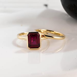 Puede incluir: Un anillo de oro con una piedra preciosa rectangular de color rojo intenso. El anillo está engastado en una banda de oro, y otra banda de oro es visible detrás. La piedra preciosa tiene una apariencia facetada, y el anillo está sobre un fondo blanco.