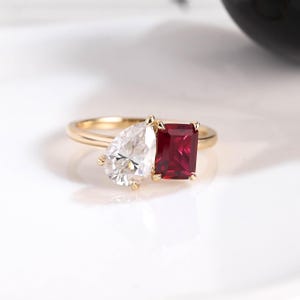 Könnte beinhalten: Ein goldener Ring mit einem birnenförmigen weißen Edelstein und einem rechteckigen roten Edelstein. Der Ring hat ein zierliches Band. Die Edelsteine sind vor einem weißen Hintergrund platziert, um ihr Funkeln hervorzuheben. Ein klassisches Schmuckstück.