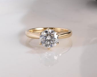 Puede incluir: Un anillo de oro con una sola piedra preciosa transparente de talla brillante redonda, sujeta por un engaste de seis garras. La banda es lisa y pulida, mostrando un diseño clásico con una superficie reflectante que resalta la claridad.