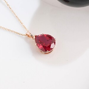May include: A rose gold necklace featuring a pear-shaped, deep red gemstone pendant. The faceted stone is secured by multiple prongs in a metallic setting, suspended from a delicate, thin rose gold chain that extends upward from the pendant.