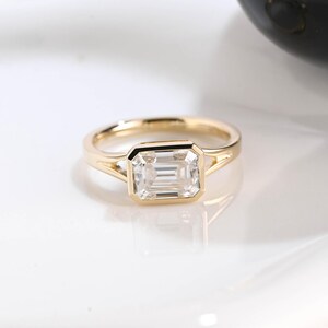 Könnte beinhalten: Ein Goldring mit einem rechteckigen, im Smaragdschliff geschliffenen, klaren Edelstein in einer Zargenfassung. Die Ringschiene besteht aus poliertem Gelbgold und weist ein geteiltes Design auf, wo sie auf die Fassung des Mittelsteins trifft.