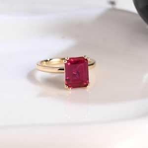 May include: A gold ring featuring a large, rectangular, vibrant red gemstone. The gemstone is the focal point, set in a four-prong setting. The ring band is a simple, polished gold. The ring is displayed on a white surface.