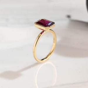 May include: A gold ring featuring a rectangular, deep red gemstone. The gemstone is set in a gold bezel, and the ring has a thin, polished gold band. The ring is displayed on a white surface.