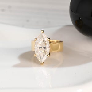 May include: A gold ring featuring a large, marquise-cut diamond. The diamond is set with four prongs and sits atop a wide, polished gold band. The ring is displayed on a white surface, reflecting light.