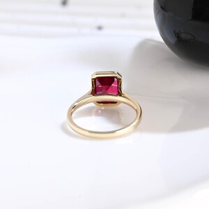Puede incluir: Un anillo de oro con una piedra preciosa roja de corte esmeralda rectangular en un engaste de bisel. La banda es lisa y pulida, mostrando un dise&ntilde;o simple y limpio que resalta el color vibrante y la forma geom&eacute;trica de la piedra central.