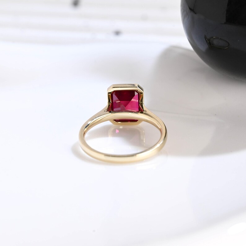 Puede incluir: Un anillo de oro con una piedra preciosa roja de corte esmeralda rectangular en un engaste de bisel. La banda es lisa y pulida, mostrando un dise&ntilde;o simple y limpio que resalta el color vibrante y la forma geom&eacute;trica de la piedra central.