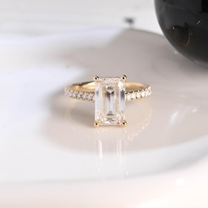 May include: A gold ring with a large, rectangular, clear gemstone as the centerpiece. The band is also gold and is encrusted with smaller, clear gemstones. The ring is set against a white background.