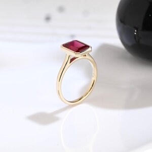 Puede incluir: Un anillo de oro con una piedra preciosa rectangular de color rojo intenso, con talla esmeralda y engastada en un bisel. La banda es fina y pulida, mostrando un acabado met&aacute;lico liso que complementa el color vibrante y saturado de la piedra.