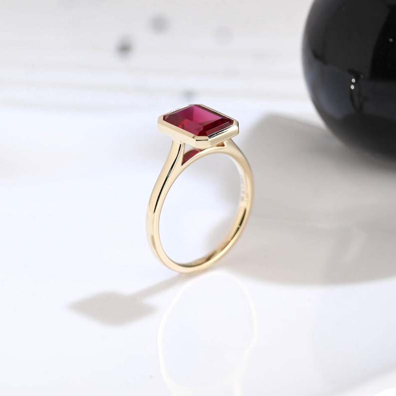 Puede incluir: Un anillo de oro con una piedra preciosa rectangular de color rojo intenso, con talla esmeralda y engastada en un bisel. La banda es fina y pulida, mostrando un acabado met&aacute;lico liso que complementa el color vibrante y saturado de la piedra.