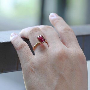 May include: A gold ring featuring a rectangular, faceted, red gemstone. The ring's band is thin and curves up to meet the gemstone setting. The gemstone is set in a bezel setting, and the ring is shown on a person's finger.