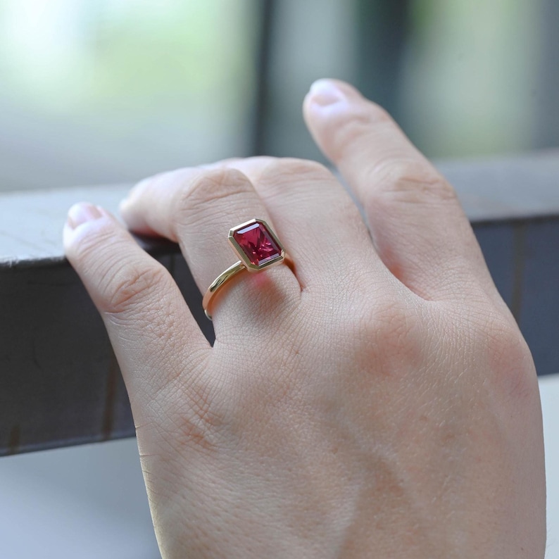 May include: A gold ring featuring a rectangular, faceted, red gemstone. The ring's band is thin and curves up to meet the gemstone setting. The gemstone is set in a bezel setting, and the ring is shown on a person's finger.