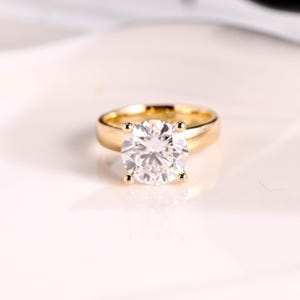 May include: A gold ring featuring a large, round, brilliant-cut clear gemstone set in a four-prong mounting. The band is smooth, polished, and solid gold, providing a classic look that highlights the central stone's clarity and reflective facets.