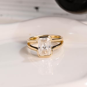 Könnte beinhalten: Ein goldener Ring mit einem rechteckigen, klaren Edelstein. Der Ring hat ein geteiltes Schaftdesign, wobei sich das Band auf beiden Seiten des Edelsteins in zwei parallele Linien aufteilt. Der Ring liegt auf einer weißen Oberfläche.
