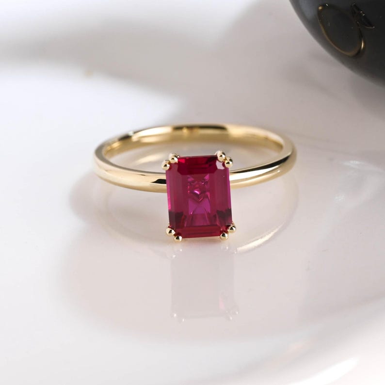 May include: A gold ring featuring a rectangular emerald-cut red gemstone set in a four-prong setting. The band is smooth and polished, showcasing a simple design that highlights the vibrant red color and faceted surface of the central stone.