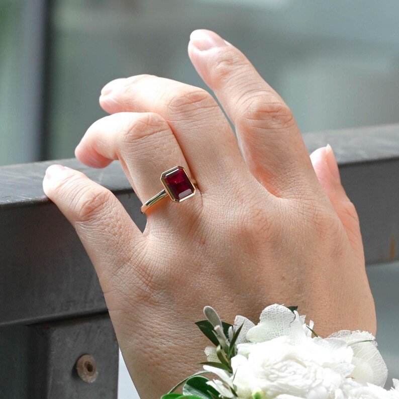 Puede incluir: Un anillo de oro con una piedra preciosa de color rojo intenso, de corte rectangular tipo esmeralda y montada en un engaste de bisel. El anillo se lleva en un dedo, mostrando la banda de oro pulido y el color vibrante de la piedra.