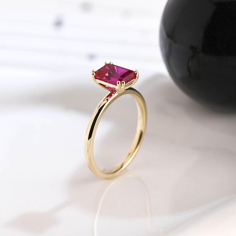 May include: A gold ring featuring a rectangular, emerald-cut vibrant pink gemstone set in a four-prong setting. The band is smooth and polished, showcasing a simple design that highlights the faceted stone and the warm metallic finish of the gold.
