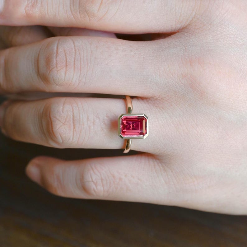 May include: A gold ring featuring a rectangular, vibrant pink gemstone. The gemstone is set in a gold bezel, creating a clean, modern look. The ring is shown on a person's finger, highlighting its elegant design and color.