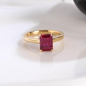 May include: A gold ring featuring a single emerald-cut, deep red gemstone set in a four-prong mounting. The band is smooth and polished, showcasing a classic design with a vibrant, saturated red stone as the central focal point of the jewelry.