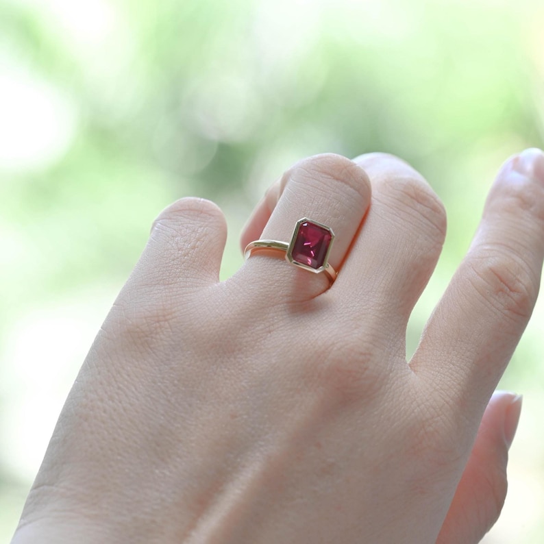 May include: A gold ring featuring a rectangular, deep red gemstone. The ring is set in a simple, elegant design, showcasing the vibrant color of the stone. The ring is on a finger, with a blurred green background.