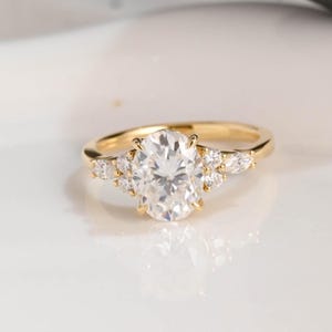May include: A gold engagement ring featuring a large oval-cut diamond as the centerpiece, flanked by smaller accent stones. The ring has a delicate band and a classic, elegant design. The diamonds sparkle against the gold setting.