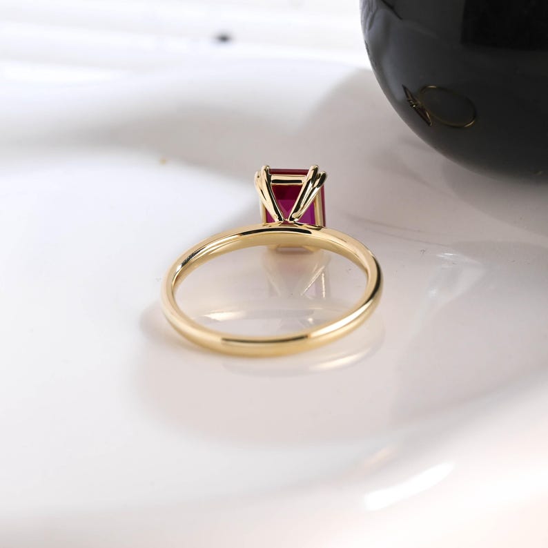 May include: A gold ring featuring a rectangular, emerald-cut vibrant red gemstone held in a four-prong setting. The band is thin, polished, and circular, showcasing a minimalist design that highlights the deep crimson color of the center stone.