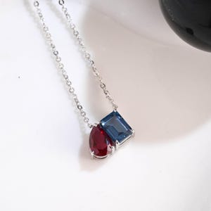 May include: A silver necklace featuring two gemstone pendants. One pendant is a deep red teardrop shape, and the other is a square, light blue gemstone. The necklace chain is delicate and the gemstones are set in a silver metal.